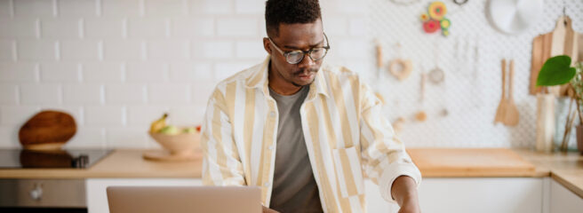 A man wearing glasses working at a table in his kitchen.