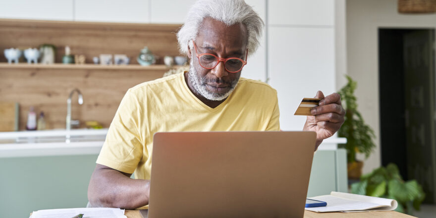 Senior man with red glasses looking at laptop with a credit card to learn about debt consolidation loan