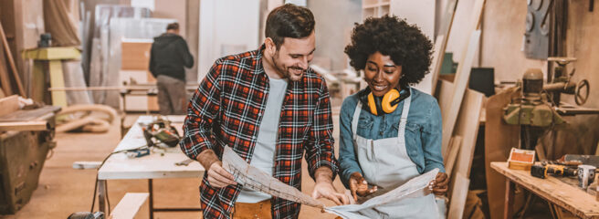 One Man and African-American woman colleagues looking at the blueprints on the paper in the interior of a modern wood workshop.