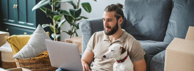 Young men moving in new house. He is happy and using laptop to organize everything. His pet dog is with him.