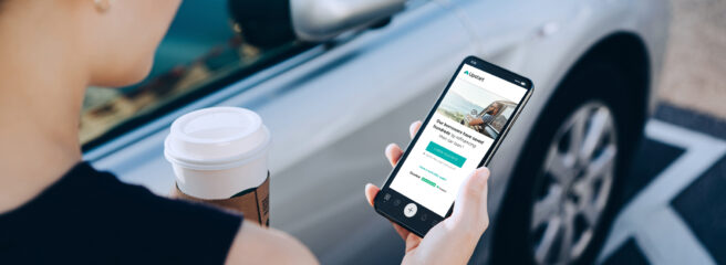 A woman holds mobile phone outside the car with a cup of coffee. Auto refinancing through Upstart appeared on the phone screen.