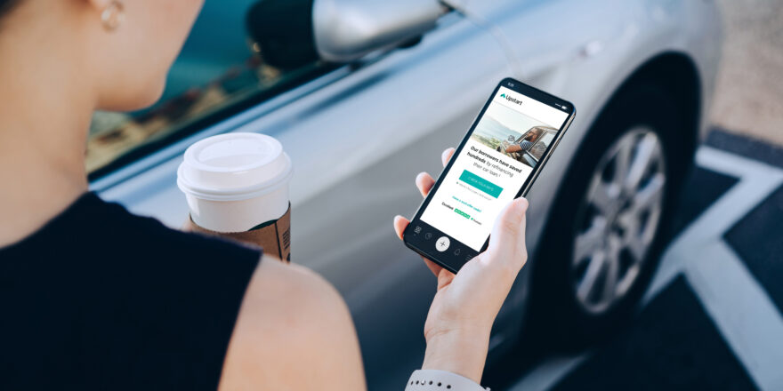 A woman holds mobile phone outside the car with a cup of coffee. Auto refinancing through Upstart appeared on the phone screen.