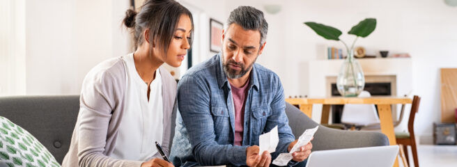 Woman with notebook making notes and man sitting next to her with receipts in hand looking at a laptop.