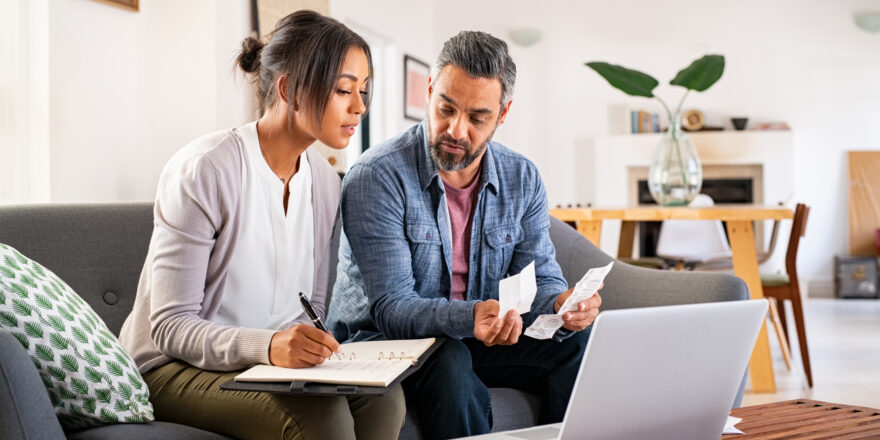 Woman with notebook making notes and man sitting next to her with receipts in hand looking at a laptop.