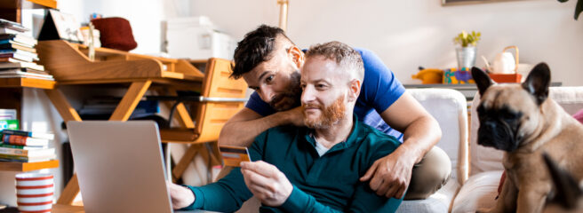 A man is sitting and looking at his laptop and holding his credit card as he works on selecting a debt consolidation company with his partner.