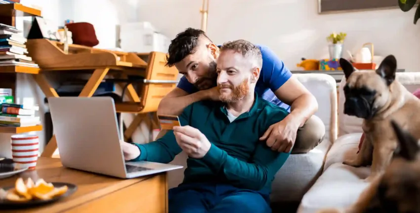 A man is sitting and looking at his laptop and holding his credit card as he works on selecting a debt consolidation company with his partner.