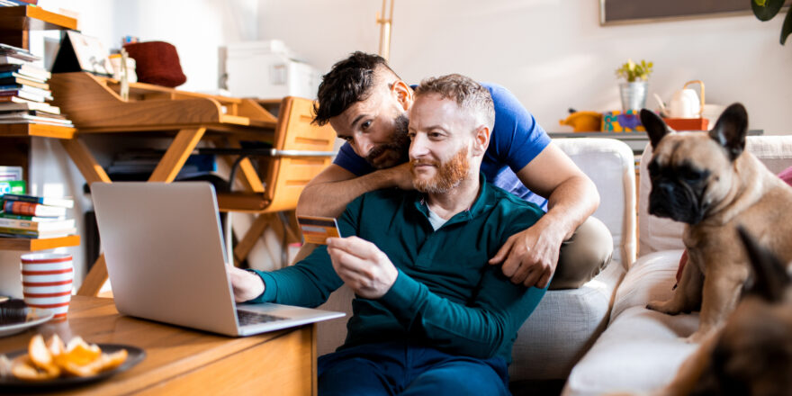 A man is sitting and looking at his laptop and holding his credit card as he works on selecting a debt consolidation company with his partner.