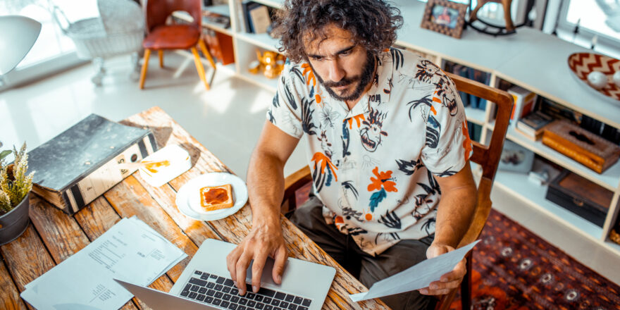 Man sitting at wood table holding a piece of paper in one hand and typing on laptop with other.