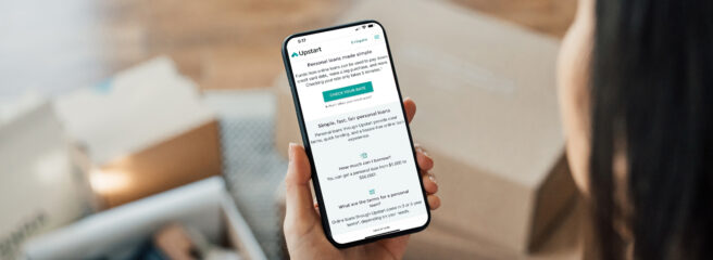 Person with dark hair facing down and looking at their iPhone at the Upstart website.