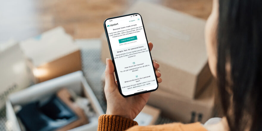 Person with dark hair facing down and looking at their iPhone at the Upstart website.
