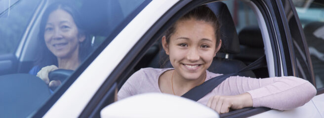A teenage girl practices driving with her mother in a white car while discussing car refinancing loans.