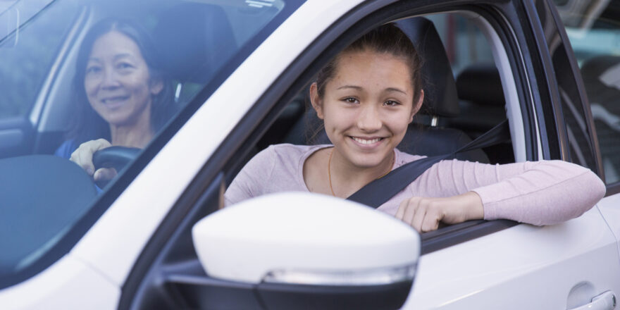 A teenage girl practices driving with her mother in a white car while discussing car refinancing loans.