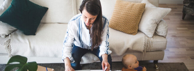 Woman sitting on a white couch working on her laptop with her toddler playing next to her.