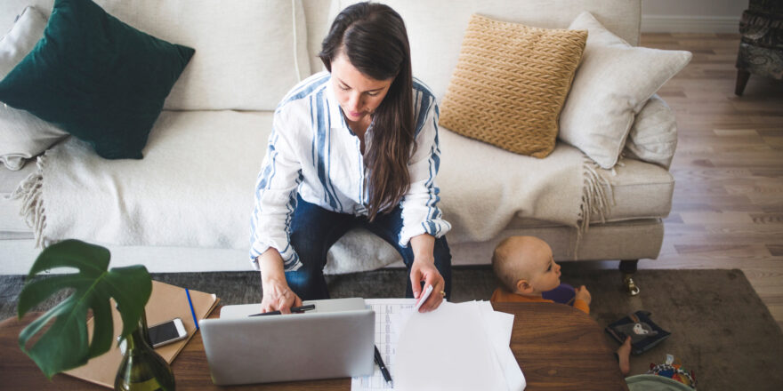 Woman sitting on a white couch working on her laptop with her toddler playing next to her.