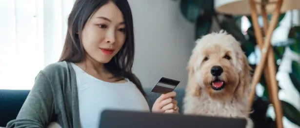 Woman sitting next to dog holding a credit card and looking at her laptop.