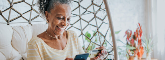 Woman is sitting at home and holding a black cell phone and credit card after she researches how to consolidate credit card debt.