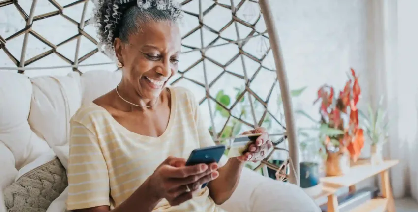 Woman is sitting at home and holding a black cell phone and credit card after she researches how to consolidate credit card debt.