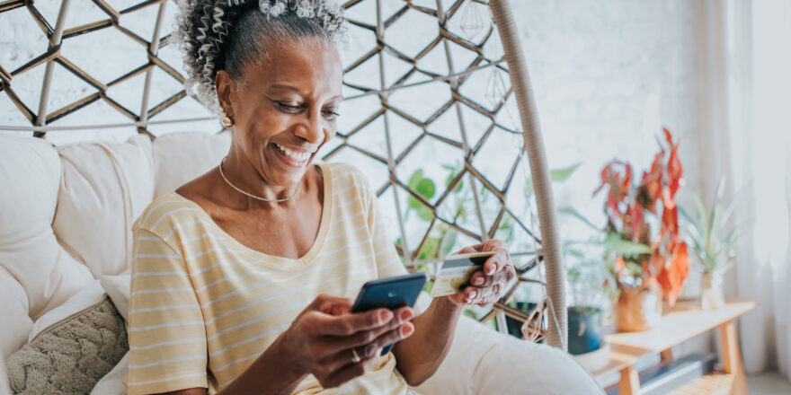 Woman is sitting at home and holding a black cell phone and credit card after she researches how to consolidate credit card debt.