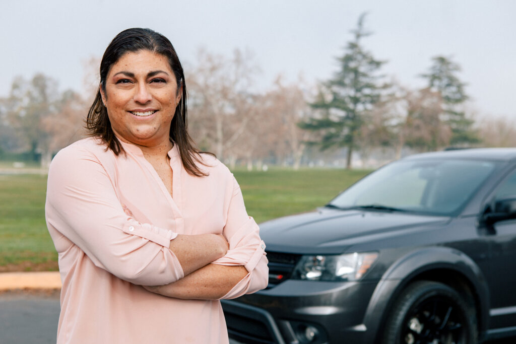 Woman in pink shirt standing outside in front of car with arms crossed.