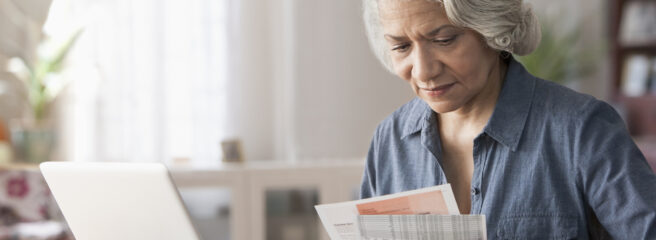 Woman in blue button down sitting at a desk looking at papers and working on her laptop.