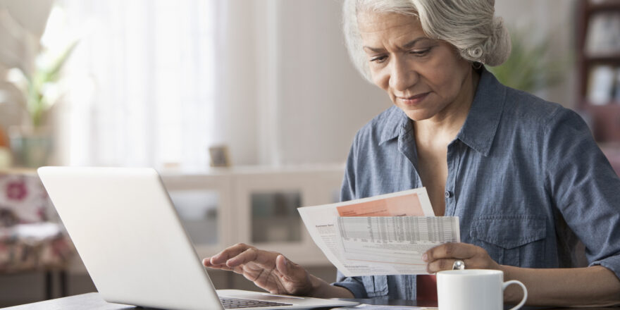 Woman in blue button down sitting at a desk looking at papers and working on her laptop.