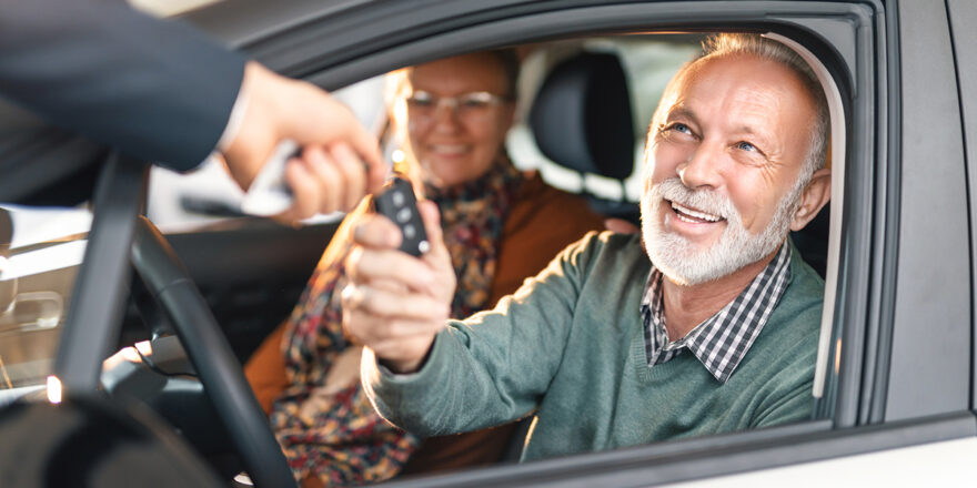 A couple sits in their new car and accepts the keys from a car salesperson