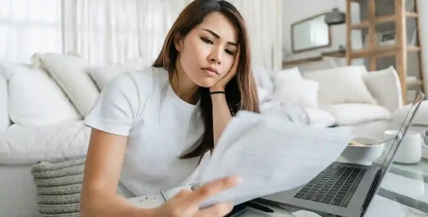 Young woman reviewing medical bills while sitting on the floor in front of her laptop