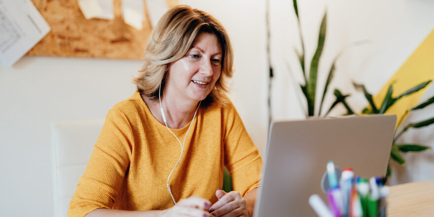 Woman in yellow shirt with headphones on and looking at her laptop.