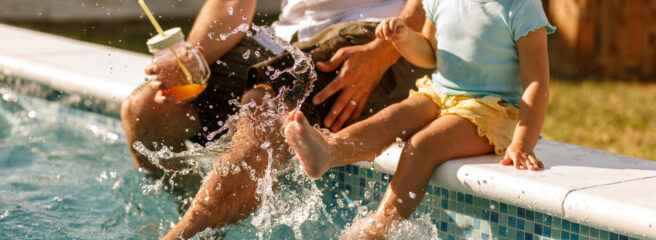 A father and daughter sit by the pool with their feet in the water on a sunny day