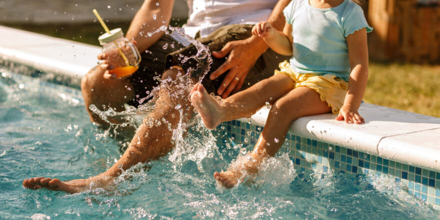 A father and daughter sit by the pool with their feet in the water on a sunny day