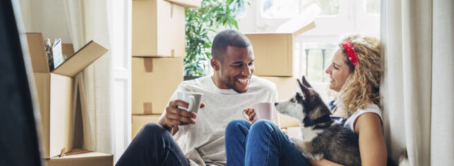 A man and woman sitting on the floor with their dog surrounded by moving boxes