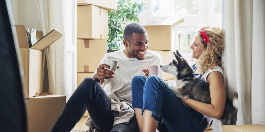 A man and woman sitting on the floor with their dog surrounded by moving boxes