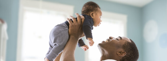 A man in a white t-shirt is smiling and holding a baby in a blue nursery.