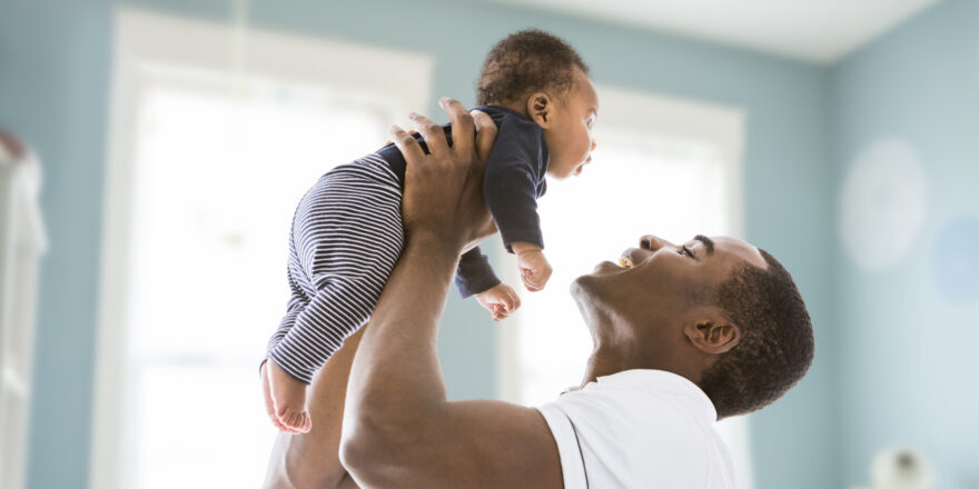 A man in a white t-shirt is smiling and holding a baby in a blue nursery.