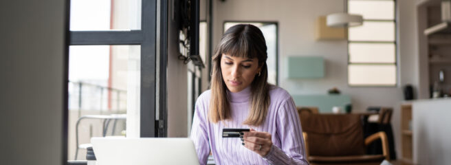 A woman sits at a table using her laptop while holding a credit card