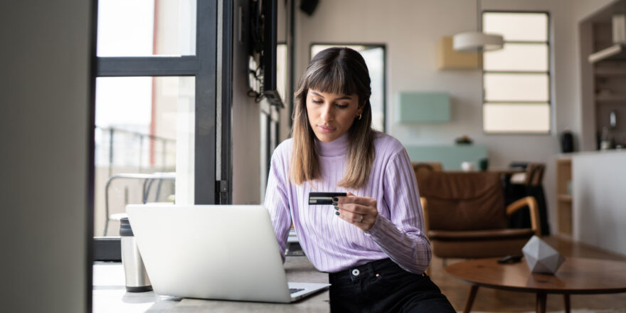 A woman sits at a table using her laptop while holding a credit card