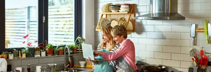 Women standing looking at a laptop in a kitchen.