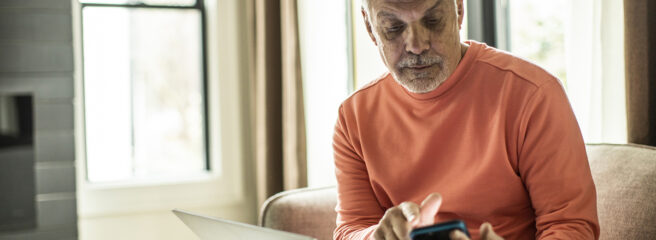 An older man researches on his phone and laptop what debt settlement is to decide if it will resolve his debt.