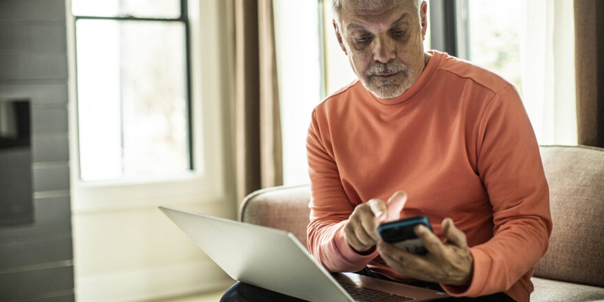 An older man researches on his phone and laptop what debt settlement is to decide if it will resolve his debt.