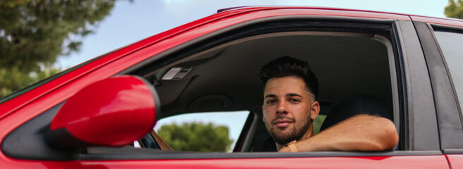 Young man inside of a red car smiling about saving on his auto refinancing loan