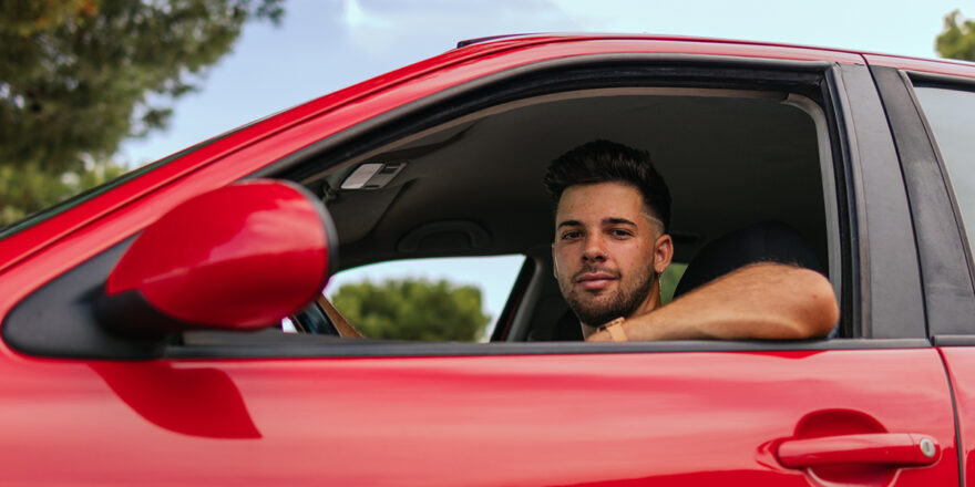 Young man inside of a red car smiling about saving on his auto refinancing loan